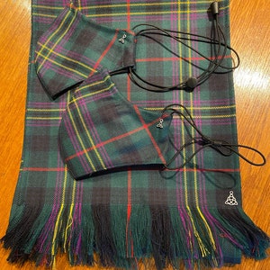 May include: A green, black, yellow and pink tartan fabric scarf with fringed edges. Two matching face masks are included with the scarf. The masks have black elastic ear loops and a small silver charm.