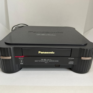 Könnte beinhalten: Eine schwarze Panasonic Real 3DO Interactive Multiplayer FZ-1 Konsole mit silberner Frontplatte und Knöpfen. Die Konsole hat einen Steckplatz für eine Spielkassette und ein CD-Laufwerk.
