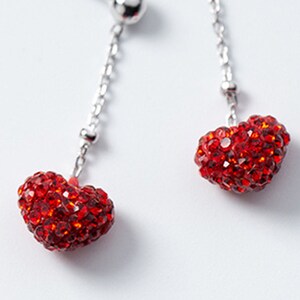 May include: A pair of drop earrings with silver posts and chains. Each earring has a small, round silver bead and a heart-shaped charm covered in red crystals. The earrings are set against a white backdrop, showcasing the jewellery's design.