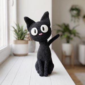 May include: A handmade black crochet cat figurine with large white eyes and a pink nose. The cat is sitting upright with one paw raised, and has long whiskers. The cat is placed on a white surface near a window.