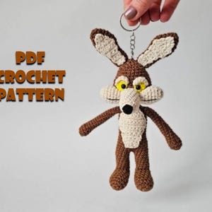 May include: A crocheted Wile E. Coyote keychain in brown, beige, and white, with yellow eyes and a black nose. The keychain is held by a silver ring. Text on the left reads "PDF CROCHET PATTERN".