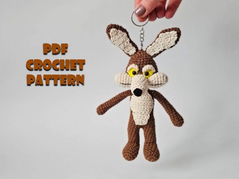 May include: A crocheted Wile E. Coyote keychain in brown, beige, and white, with yellow eyes and a black nose. The keychain is held by a silver ring. Text on the left reads "PDF CROCHET PATTERN".