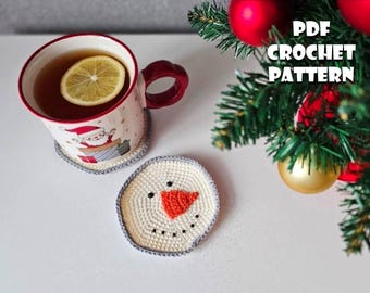 Könnte beinhalten: Eine festliche Szene mit einer Tasse mit einer Zitronenscheibe, die auf einem gehäkelten Untersetzer steht. Ein Schneemann-Untersetzer liegt daneben. Die Tasse hat ein Weihnachtsmann-Design. Der Text "PDF CROCHET PATTERN" ist sichtbar. Weihnachtsbaumschmuck im Hintergrund.