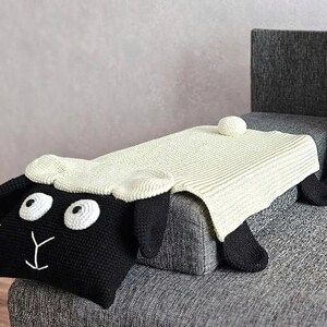 May include: A handmade, black and white sheep-shaped pillow with a cream-colored blanket draped over it. The sheep has large white eyes, a cream-colored head, and black ears and legs. The blanket is a light cream color and has a ribbed texture.