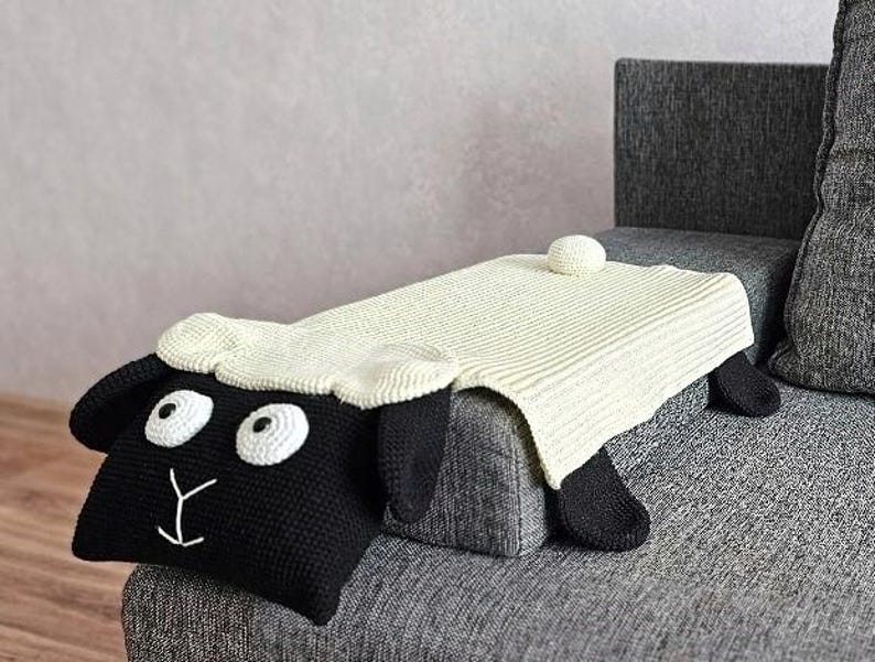 May include: A handmade, black and white sheep-shaped pillow with a cream-colored blanket draped over it. The sheep has large white eyes, a cream-colored head, and black ears and legs. The blanket is a light cream color and has a ribbed texture.