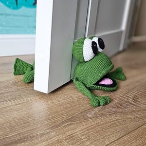 May include: A green crocheted frog doorstop with large white eyes and a pink open mouth. The frog is positioned against a white door.