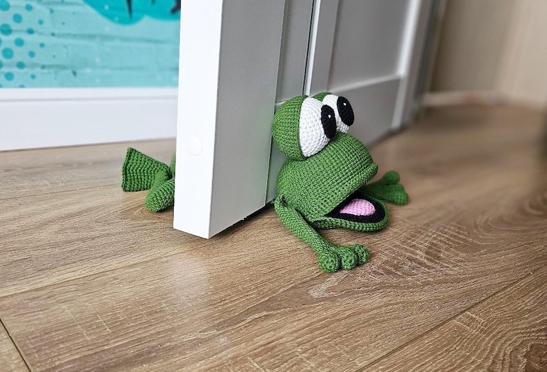 May include: A green crocheted frog doorstop with large white eyes and a pink open mouth. The frog is positioned against a white door.