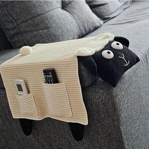 May include: A cream-colored knitted sheep-shaped couch armrest organizer with two pockets. The sheep has a black head with white eyes and a smiling face. Black legs extend down the side of the couch. Two remote controls are in the pockets.