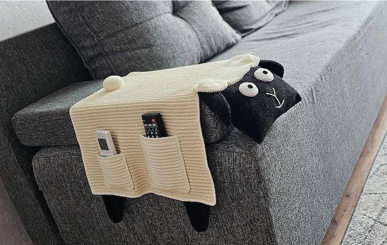 May include: A cream-colored knitted sheep-shaped couch armrest organizer with two pockets. The sheep has a black head with white eyes and a smiling face. Black legs extend down the side of the couch. Two remote controls are in the pockets.