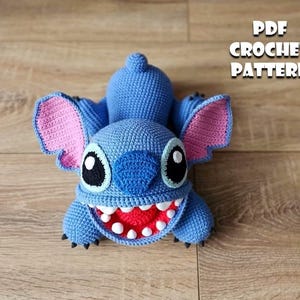 Op de afbeelding: Een blauwe Stitch-pop gehaakt met grote oren, een brede grijns en zwarte ogen. De pop heeft een getextureerd uiterlijk en ligt op een houten oppervlak. Tekst rechts zegt "PDF CROCHET PATTERN".