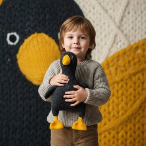 Könnte beinhalten: Ein Kind hält ein dunkelgraues, gestricktes Gänse-Plüschtier mit gelben Füßen und Schnabel. Die Gans hat einen freundlichen Ausdruck. Der Hintergrund zeigt einen großen, gestrickten Gänsekopf in Dunkelgrau und Gelb sowie eine cremefarbene und gelbe Strickdecke.