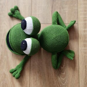 May include: Two green crocheted frog toys. One frog has large white eyes with black pupils and a wide open mouth. The other frog has a round body and small eyes.