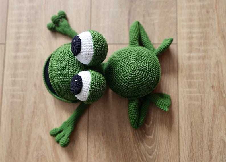 May include: Two green crocheted frog toys. One frog has large white eyes with black pupils and a wide open mouth. The other frog has a round body and small eyes.