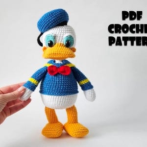 Op de afbeelding: Een gehaakte Donald Duck pop, met een blauw matrozenhemd, hoed en vlinderdas. De pop heeft een wit gezicht, gele snavel en voeten en zwarte ogen. De tekst "PDF CROCHET PATTERN" wordt in het zwart weergegeven.