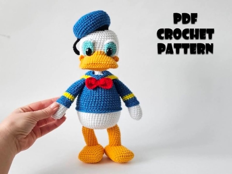 Op de afbeelding: Een gehaakte Donald Duck pop, met een blauw matrozenhemd, hoed en vlinderdas. De pop heeft een wit gezicht, gele snavel en voeten en zwarte ogen. De tekst "PDF CROCHET PATTERN" wordt in het zwart weergegeven.