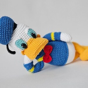 Op de afbeelding: Gehaakte Donald Duck pop in liggende positie. De pop heeft een blauwe matrozenmuts, een wit gezicht en een gele snavel. Het heeft een blauw lichaam met een witte buik, gele voeten en een rode strik. Handgemaakt.