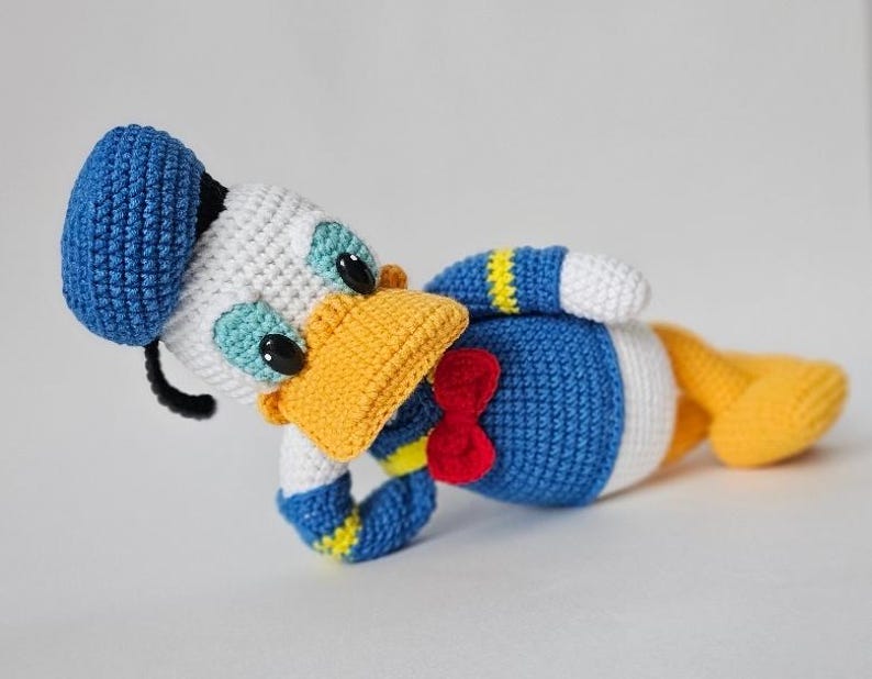 Op de afbeelding: Gehaakte Donald Duck pop in liggende positie. De pop heeft een blauwe matrozenmuts, een wit gezicht en een gele snavel. Het heeft een blauw lichaam met een witte buik, gele voeten en een rode strik. Handgemaakt.