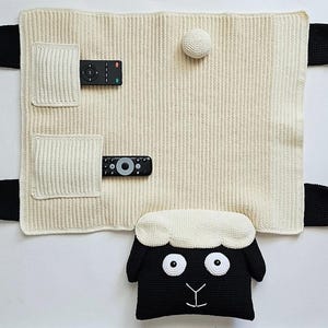 May include: A cream-colored, knitted TV remote control organizer shaped like a sheep. The organizer has two pockets for remotes, black ears, and a sheep head pillow at the bottom. A white ball is attached to the top.