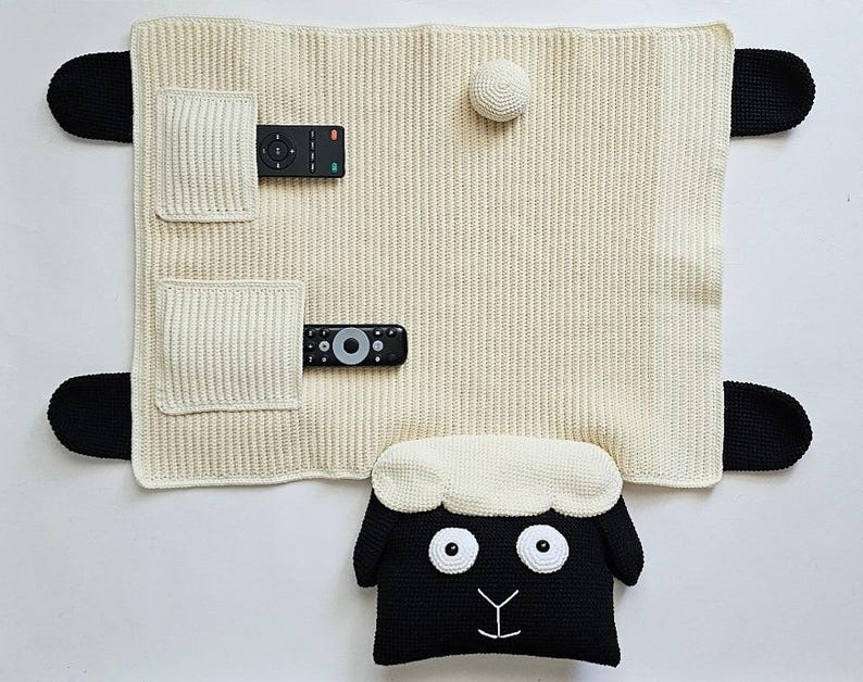 May include: A cream-colored, knitted TV remote control organizer shaped like a sheep. The organizer has two pockets for remotes, black ears, and a sheep head pillow at the bottom. A white ball is attached to the top.