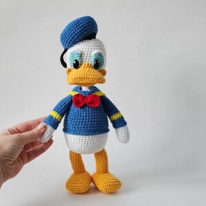 Op de afbeelding: Een handgemaakte gehaakte Donald Duck pop. De pop heeft een blauw matrozenhemd, een rode vlinderdas en een blauwe matrozenmuts. De snavel en voeten zijn geel en de ogen zijn blauw. De pop is ongeveer 25 cm hoog.