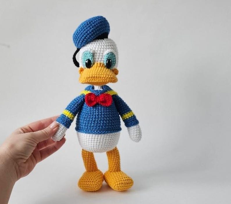 Op de afbeelding: Een handgemaakte gehaakte Donald Duck pop. De pop heeft een blauw matrozenhemd, een rode vlinderdas en een blauwe matrozenmuts. De snavel en voeten zijn geel en de ogen zijn blauw. De pop is ongeveer 25 cm hoog.