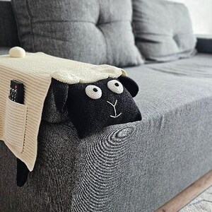 May include: Cream-colored knitted sofa armrest cover with two pockets, holding a remote and a phone. A black sheep head with white eyes and a white embroidered smile is attached to the cover. The cover is on a gray sofa.