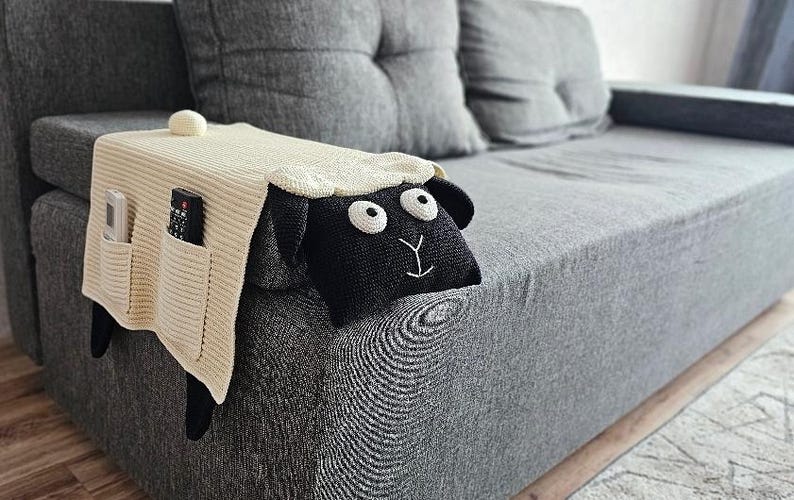 May include: Cream-colored knitted sofa armrest cover with two pockets, holding a remote and a phone. A black sheep head with white eyes and a white embroidered smile is attached to the cover. The cover is on a gray sofa.