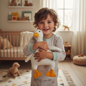 May include: A child in pyjamas holds a white knitted goose toy with yellow feet and beak. The child is smiling. The background includes a cot, a stuffed dog, and a window.