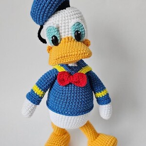 Op de afbeelding: Een handgemaakte gehaakte Donald Duck-pop. De pop heeft een blauw matrozenhemd met gele accenten, een rode vlinderdas en een blauwe matrozenmuts. De eend heeft een gele snavel en voeten, een wit gezicht en buik en zwarte ogen.