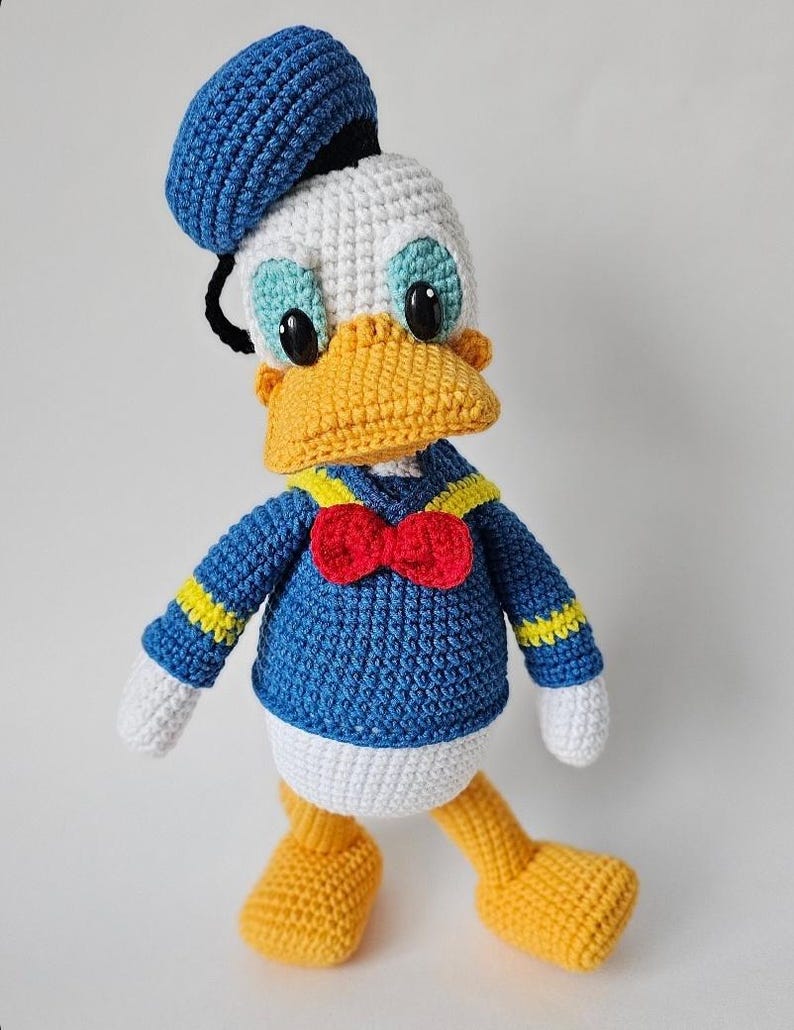 Op de afbeelding: Een handgemaakte gehaakte Donald Duck-pop. De pop heeft een blauw matrozenhemd met gele accenten, een rode vlinderdas en een blauwe matrozenmuts. De eend heeft een gele snavel en voeten, een wit gezicht en buik en zwarte ogen.