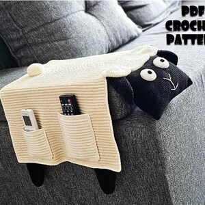 May include: A cream-colored crochet sheep-shaped couch armrest organizer with two pockets, holding a remote and a phone. The sheep has a black face with white eyes and a white woolly top. The text "PDF CROCHET PATTERN" is visible.