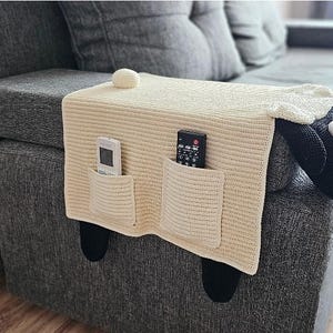 May include: Cream-colored knitted sofa armrest cover shaped like a sheep, with two pockets holding a remote control and a TV remote. The sheep has a black face, white wool, and black legs. A decorative ball sits on top.