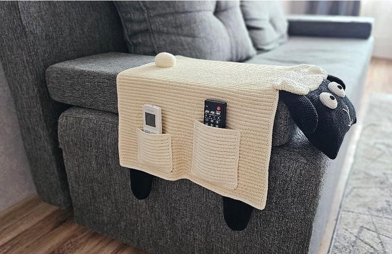 May include: Cream-colored knitted sofa armrest cover shaped like a sheep, with two pockets holding a remote control and a TV remote. The sheep has a black face, white wool, and black legs. A decorative ball sits on top.