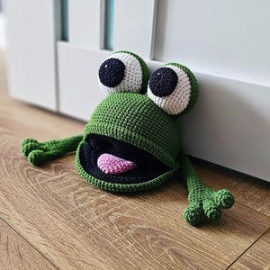 May include: Green crocheted frog doorstop with black eyes, white pupils, and a pink tongue. The frog is sitting on a wooden floor.