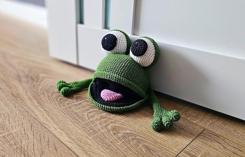 May include: Green crocheted frog doorstop with black eyes, white pupils, and a pink tongue. The frog is sitting on a wooden floor.