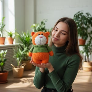 May include: A handmade orange teddy bear with a white face and a green jumper. The bear has small ears and a pink nose. The plush toy is held in front of a background of green plants.