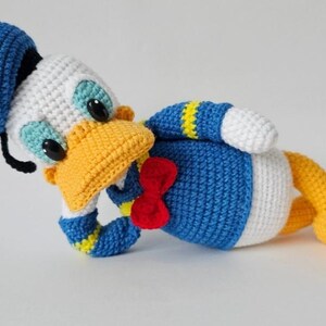 Op de afbeelding: Gehaakte Donald Duck pluche speelgoed. Het speelgoed heeft een blauwe matrozenmuts, blauw shirt, witte buik, gele voeten en een gele snavel. Het heeft een rode vlinderdas en zwarte ogen. Het pluche ligt op zijn zij.