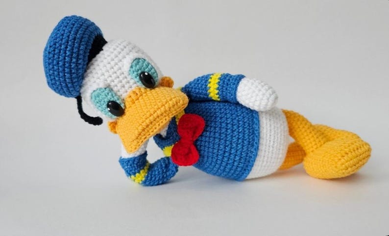 Op de afbeelding: Gehaakte Donald Duck pluche speelgoed. Het speelgoed heeft een blauwe matrozenmuts, blauw shirt, witte buik, gele voeten en een gele snavel. Het heeft een rode vlinderdas en zwarte ogen. Het pluche ligt op zijn zij.
