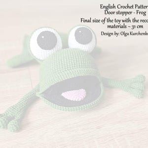 May include: Green crocheted frog doorstop with large white eyes and a pink tongue. The frog is 31 cm in size.