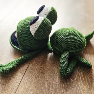 May include: A green crocheted frog toy with large eyes and a long tail. The frog is lying on its back with its legs in the air. There is a smaller green crocheted turtle toy next to the frog.