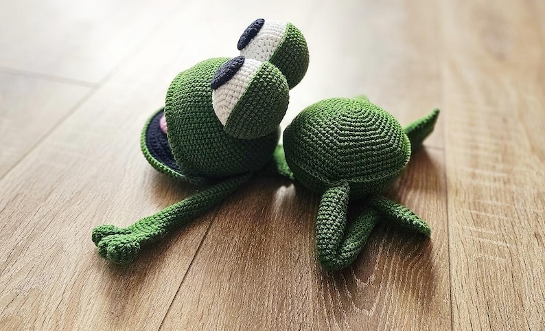 May include: A green crocheted frog toy with large eyes and a long tail. The frog is lying on its back with its legs in the air. There is a smaller green crocheted turtle toy next to the frog.