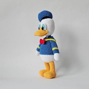 Op de afbeelding: Gehaakte Donald Duck pop. Het handgemaakte speelgoed heeft een blauw matrozenhemd met gele biezen, een rode vlinderdas en een blauwe matrozenmuts. De eend heeft een witte kop, een gele snavel en gele voeten. De pop staat.