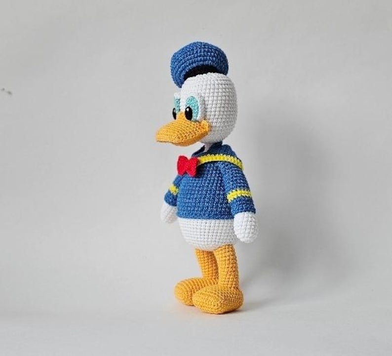 Op de afbeelding: Gehaakte Donald Duck pop. Het handgemaakte speelgoed heeft een blauw matrozenhemd met gele biezen, een rode vlinderdas en een blauwe matrozenmuts. De eend heeft een witte kop, een gele snavel en gele voeten. De pop staat.