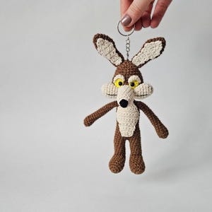 May include: Crocheted keychain featuring a cartoon coyote character. The coyote has brown fur, cream-colored accents, and yellow eyes. It is attached to a silver-colored keyring. The keychain is handmade.