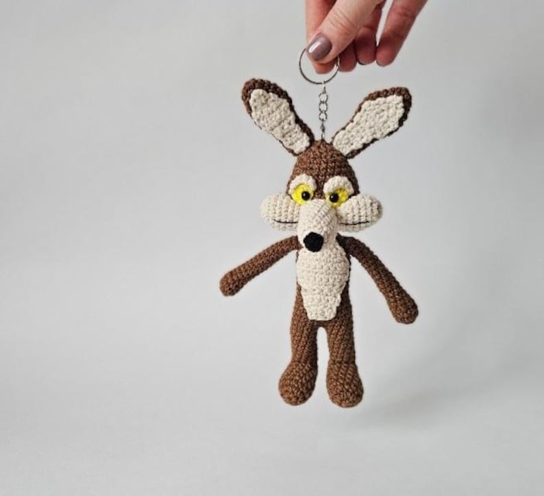 May include: Crocheted keychain featuring a cartoon coyote character. The coyote has brown fur, cream-colored accents, and yellow eyes. It is attached to a silver-colored keyring. The keychain is handmade.