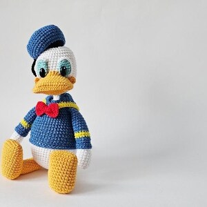 Op de afbeelding: Een gehaakte Donald Duck pop. De pop draagt een blauw matrozenhemd met gele strepen, een rode vlinderdas en een blauwe matrozenmuts. Het heeft een gele snavel en voeten, en een wit gezicht en handen.