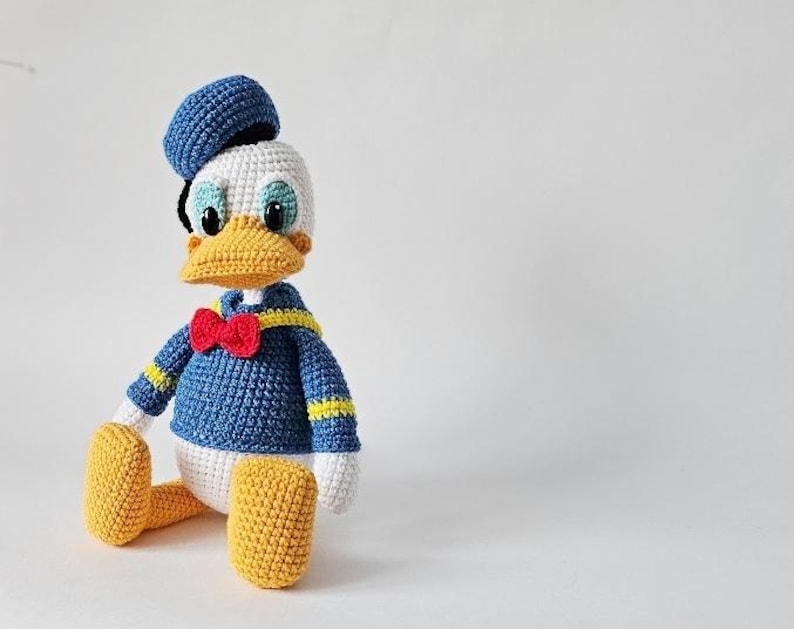 Op de afbeelding: Een gehaakte Donald Duck pop. De pop draagt een blauw matrozenhemd met gele strepen, een rode vlinderdas en een blauwe matrozenmuts. Het heeft een gele snavel en voeten, en een wit gezicht en handen.
