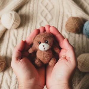 May include: A small, brown knitted teddy bear with a white muzzle and black eyes is held in cupped hands. The bear is surrounded by balls of yarn in various neutral colors, set against a cream-colored knitted background.