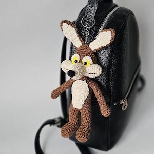 May include: A handmade crochet keychain featuring a cartoon coyote character with brown, beige, and black details. The keychain is attached to a black backpack. The coyote has large ears, yellow eyes, and a white chest.