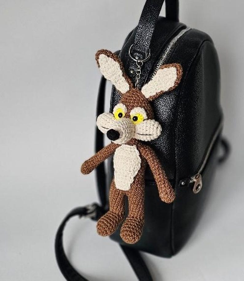 May include: A handmade crochet keychain featuring a cartoon coyote character with brown, beige, and black details. The keychain is attached to a black backpack. The coyote has large ears, yellow eyes, and a white chest.